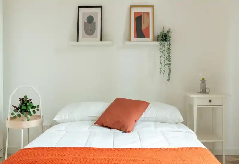 Camera da letto luminosa e minimal con letto ben rifatto, copriletto e cuscino arancione, tavolini semplici e quadri: uno spazio tranquillo e accogliente.