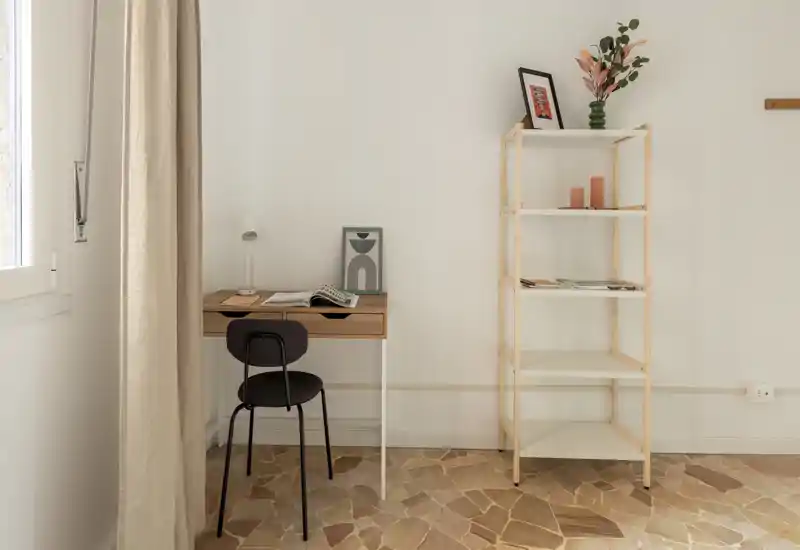 Spazio living minimal e luminoso con una piccola scrivania, sedia e scaffale aperto; tonalità neutre e luce naturale creano un angolo di lavoro tranquillo.