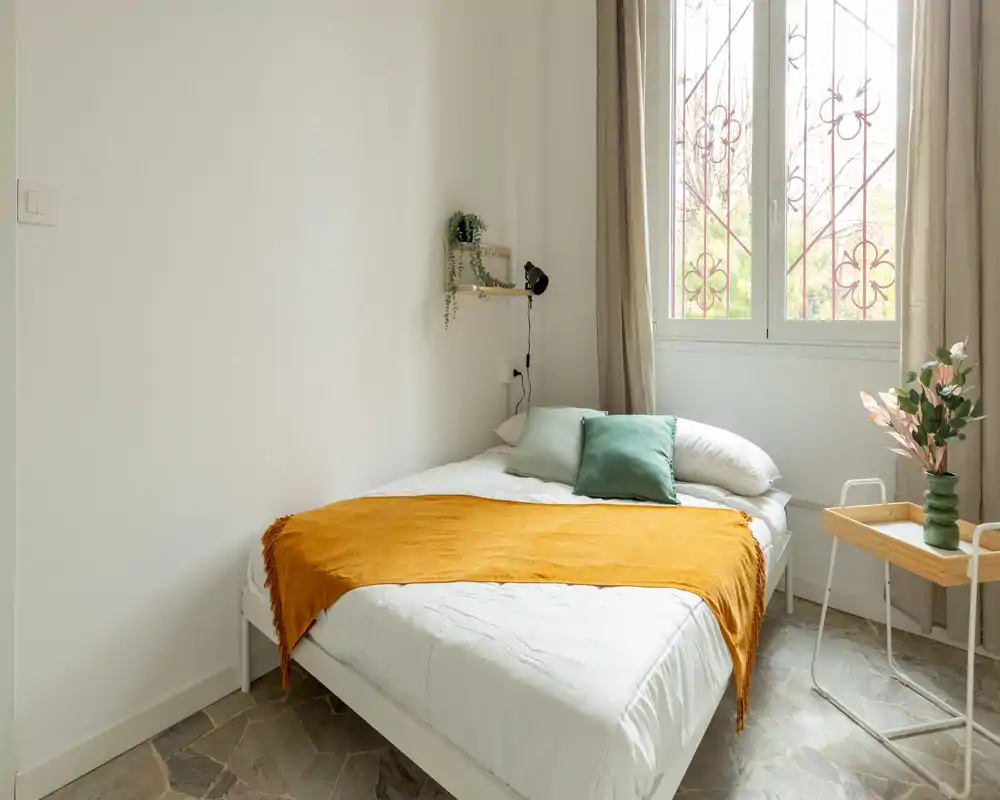 Camera da letto luminosa e curata con un comodo letto matrimoniale, plaid ocra caldo e luce naturale da una grande finestra.