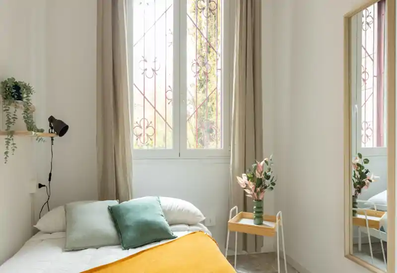 Camera da letto luminosa e accogliente con letto ben rifatto, cuscini morbidi e una coperta color senape vicino a una grande finestra; piante e uno specchio intero valorizzano l’ambiente.