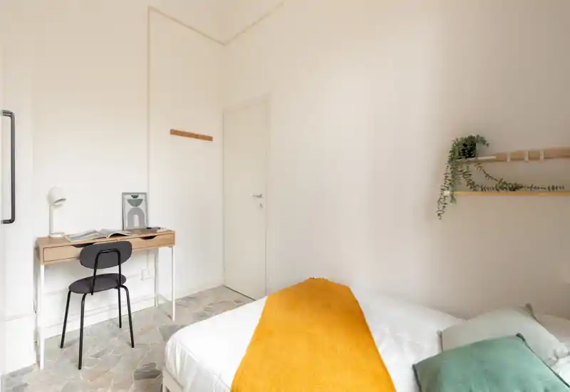 Camera da letto luminosa e minimal con letto ordinato e una coperta color senape, una scrivania compatta e una mensola con piante — ideale per un singolo o ospiti.
