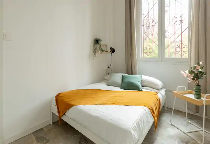 Camera da letto luminosa e curata con un comodo letto matrimoniale, plaid ocra caldo e luce naturale da una grande finestra.