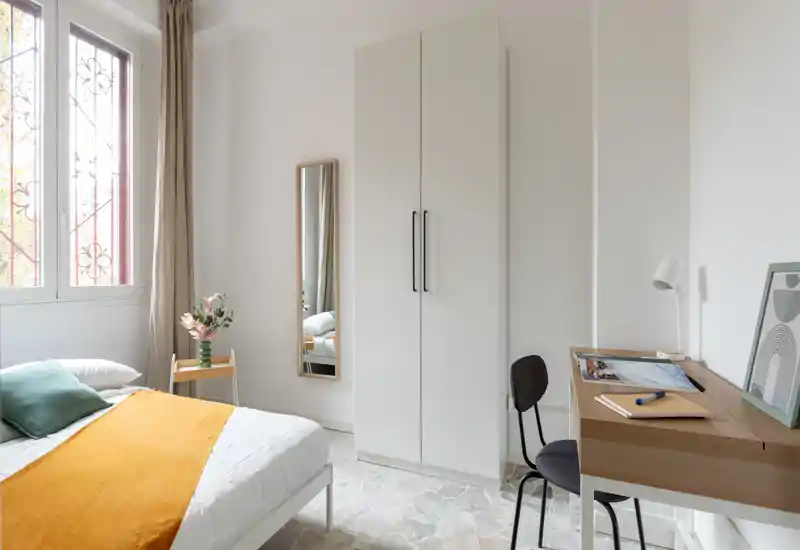 Camera da letto luminosa e minimalista con letto matrimoniale in lenzuola bianche e un caldo copriletto arancione; presenta un armadio alto, uno specchio a figura intera e una scrivania con sedia vicino alla finestra, spazio funzionale e tranquillo.