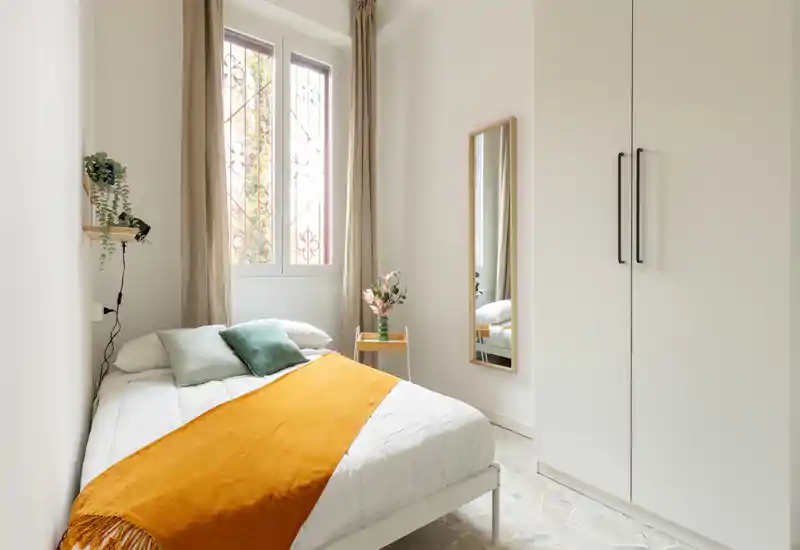 Camera da letto compatta e luminosa con letto ben rifatto, plaid arancione caldo, cuscini verdi e armadio alto. La luce naturale dalla finestra e l’arredo semplice e moderno creano un’atmosfera tranquilla e accogliente.
