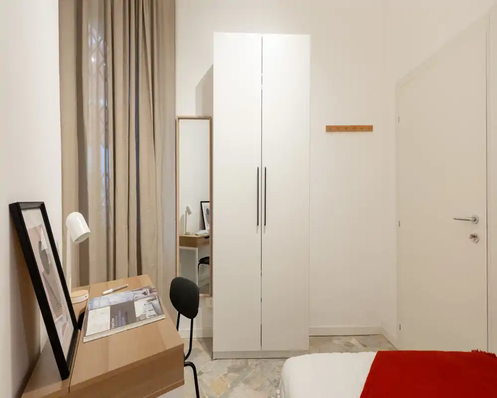 Angolo camera da letto moderno e ordinato con bordo del letto visibile, armadio bianco, scrivania e sedia — luminoso, minimal e ben curato per un soggiorno comodo.