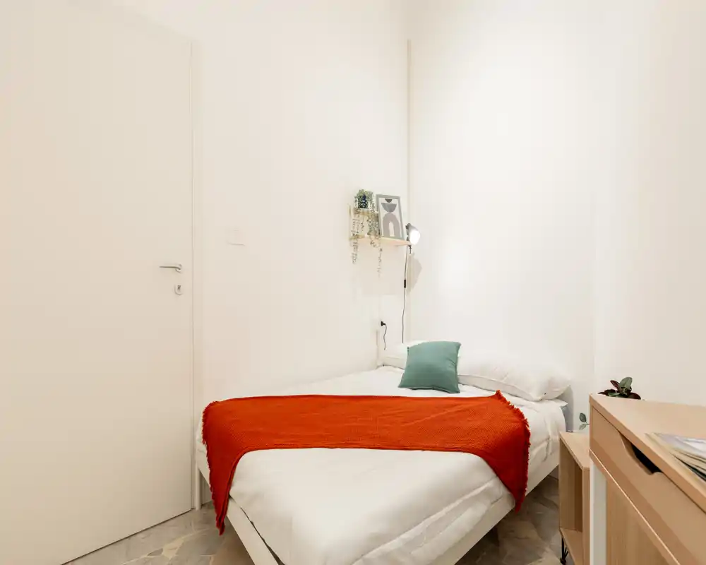 Camera da letto singola minimal e luminosa con letto ben rifatto, plaid arancione e cuscino verde; piccola mensola e lampada da comodino creano un ambiente pulito e accogliente, perfetto per affitti brevi.