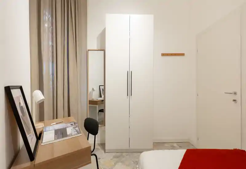Angolo camera da letto moderno e ordinato con bordo del letto visibile, armadio bianco, scrivania e sedia — luminoso, minimal e ben curato per un soggiorno comodo.
