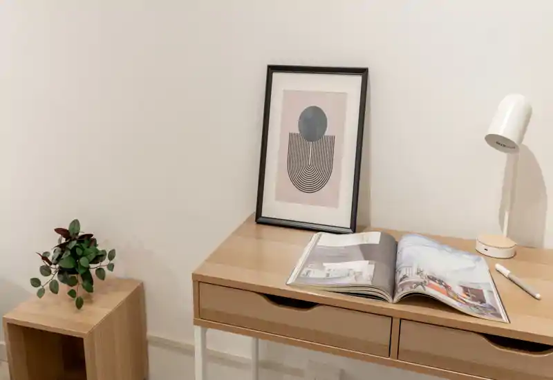 Un angolo di lavoro minimalista con scrivania in legno chiaro, lampada da tavolo, quadro incorniciato e una piccola pianta — luminoso, pulito e ben arredato per un soggiorno compatto o un angolo home office.