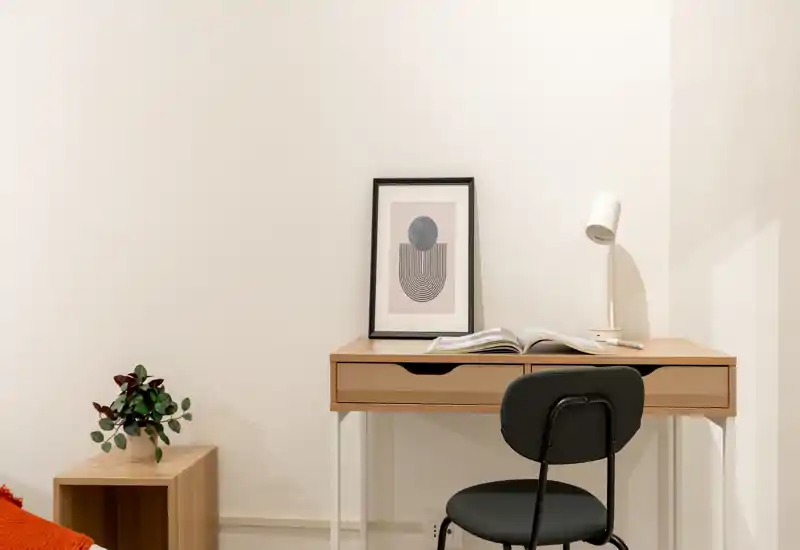 Angolo camera minimalista con scrivania in legno, lampada da tavolo, quadro incorniciato e sedia — luminoso, ordinato e adatto come piccolo spazio di lavoro in camera.