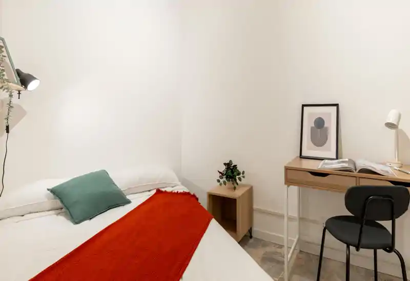 Camera da letto luminosa e minimal con letto matrimoniale in lenzuola bianche, coperta rossa e cuscino verde; piccolo scrittoio e sedia laterali per un angolo lavoro compatto.