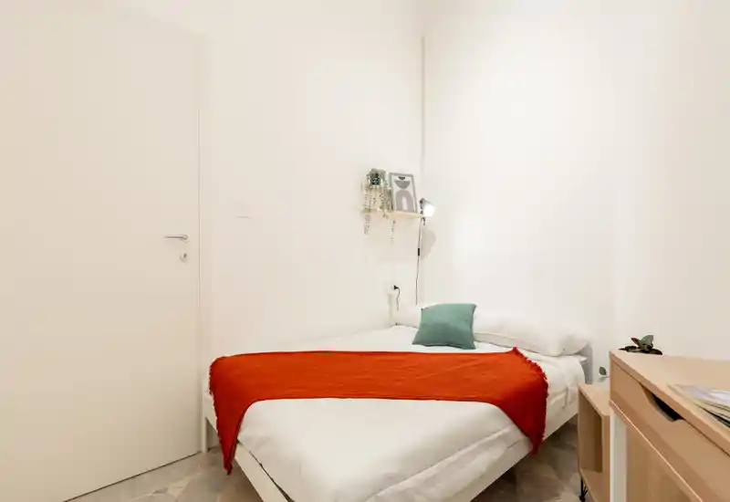 Camera da letto singola minimal e luminosa con letto ben rifatto, plaid arancione e cuscino verde; piccola mensola e lampada da comodino creano un ambiente pulito e accogliente, perfetto per affitti brevi.