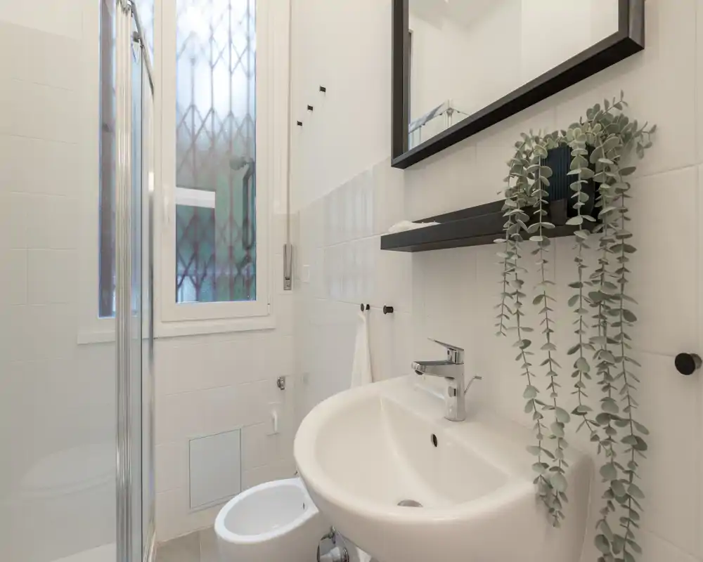 Bagno compatto e luminoso con lavabo a parete, bidet e porta doccia in vetro. Piastrelle bianche pulite e una mensola specchio donano un aspetto moderno e fresco.