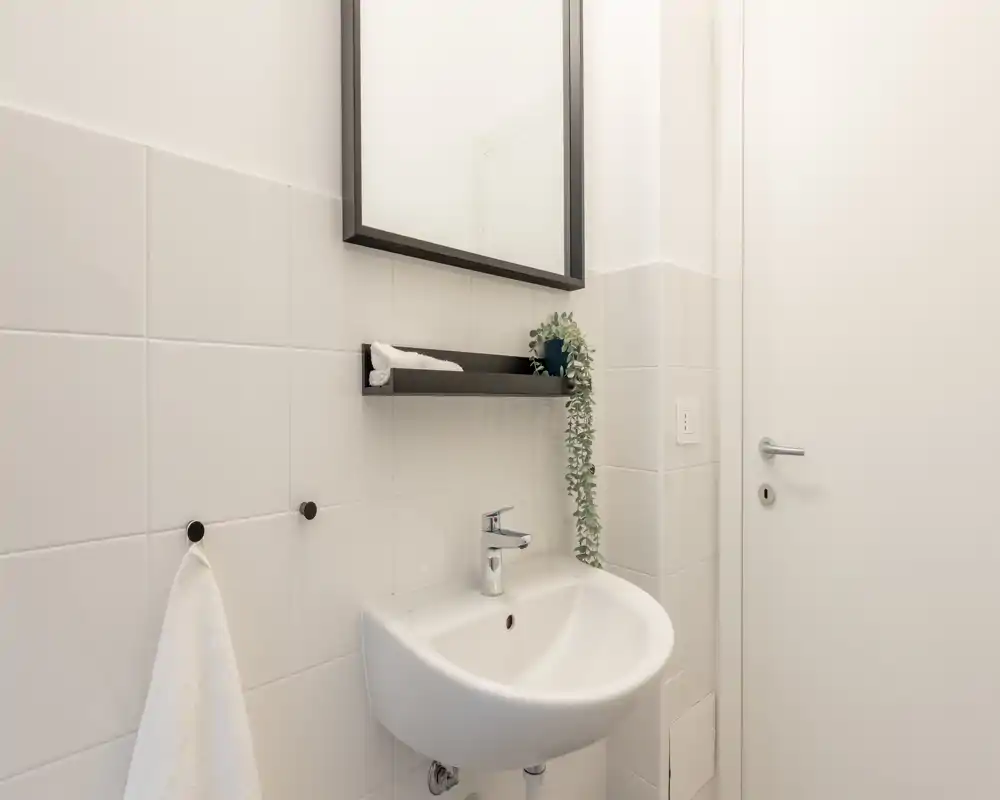 Piccolo lavabo bagno moderno con specchio a parete, mensola e asciugamano; piastrelle bianche pulite e accessori minimalisti.