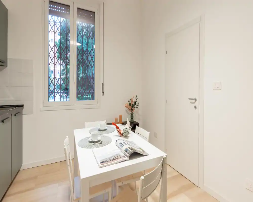 Zona pranzo ordinata con un tavolo bianco apparecchiato, sedie e dettagli decorativi vicino a una finestra luminosa — uno spazio compatto e moderno ideale per i pasti.