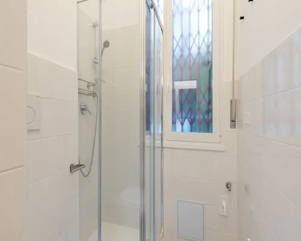 Bagno compatto e moderno con doccia con pareti in vetro, sanitari a parete e finestra opaca che offre luce naturale garantendo privacy.