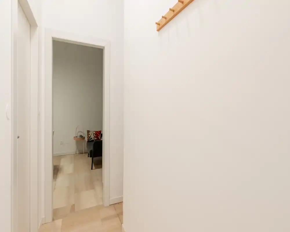 Ingresso compatto e pulito con pareti chiare, pavimento piastrellato e appendiabiti in legno; tramite la porta si intravede il soggiorno sullo sfondo.