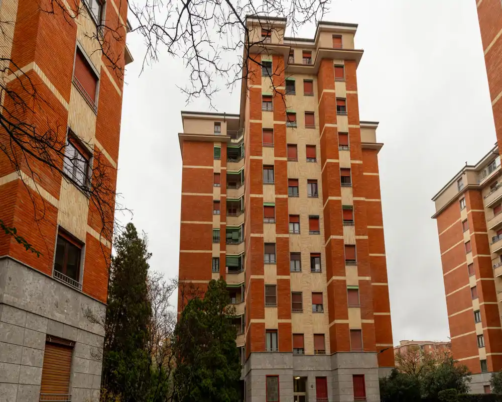 Vista esterna di un edificio residenziale multipiano con facciate in mattone rosso e cemento, cortile con alberi e cielo nuvoloso.