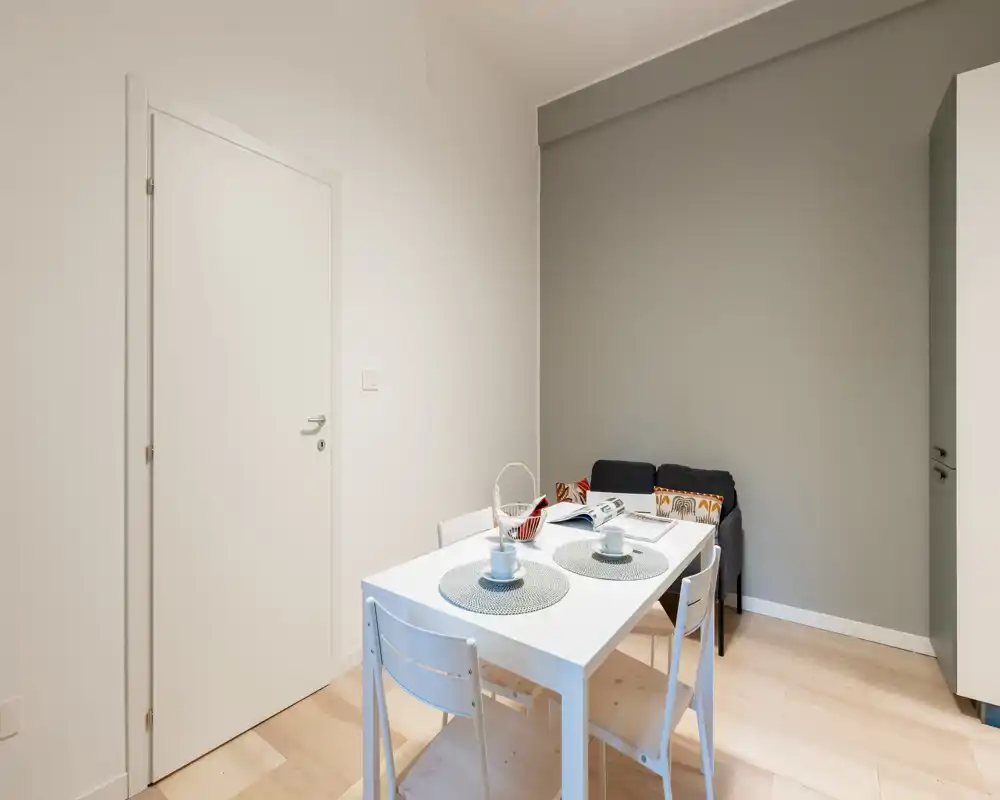 Zona pranzo luminosa e minimale con tavolo bianco apparecchiato per due e sedie semplici, adiacente a un piccolo angolo salotto; stile moderno e pulito ideale per appartamenti compatti.