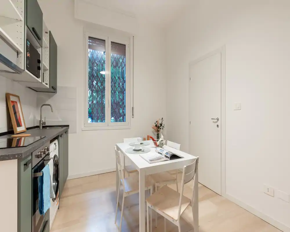 Cucina moderna e luminosa con elettrodomestici integrati e un piccolo tavolo da pranzo per quattro persone, arredata con una pianta e stoviglie per un’atmosfera accogliente.