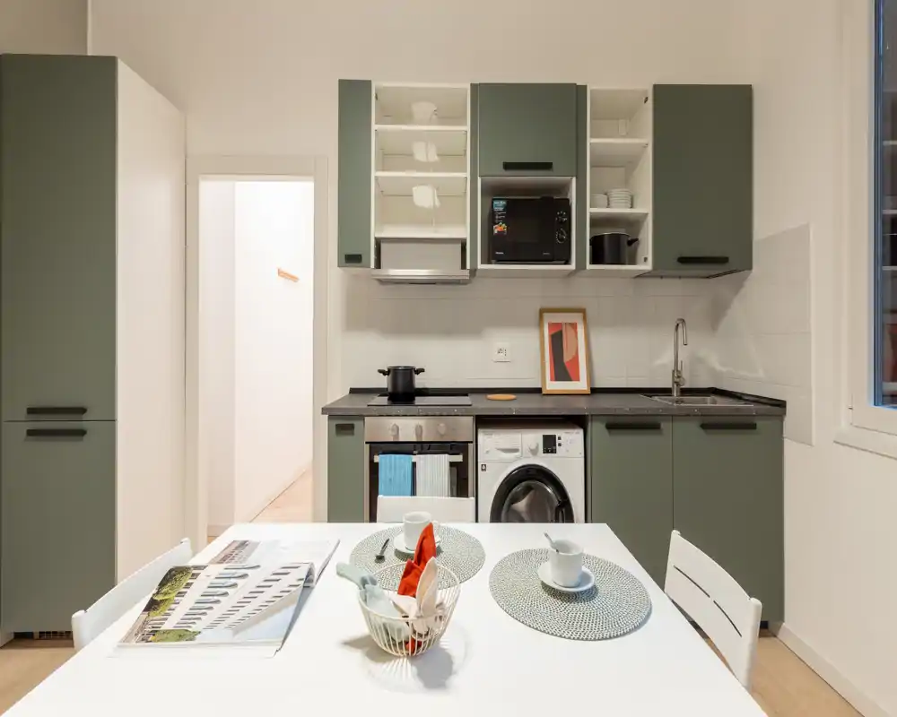 Cucina moderna compatta con pensili verdi, elettrodomestici integrati e un piccolo tavolo da pranzo per due — luminosa, ordinata e ideale per la vita in città.