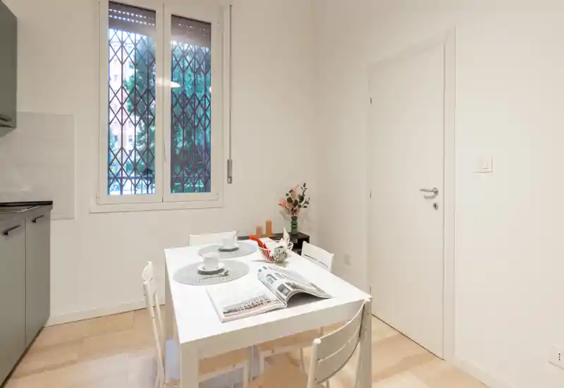 Zona pranzo ordinata con un tavolo bianco apparecchiato, sedie e dettagli decorativi vicino a una finestra luminosa — uno spazio compatto e moderno ideale per i pasti.