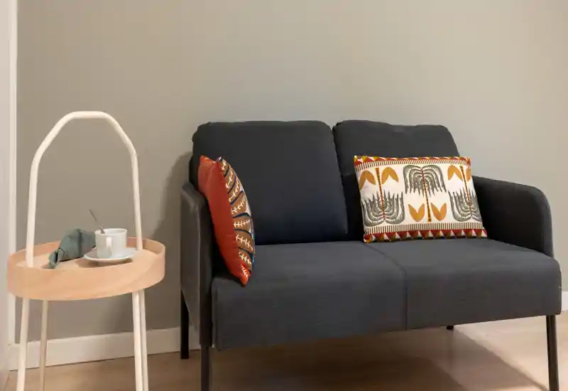 Un divano due posti elegante e accogliente con cuscini decorativi su una parete neutra, abbinato a un piccolo tavolino rotondo — un angolo soggiorno tranquillo e invitante.