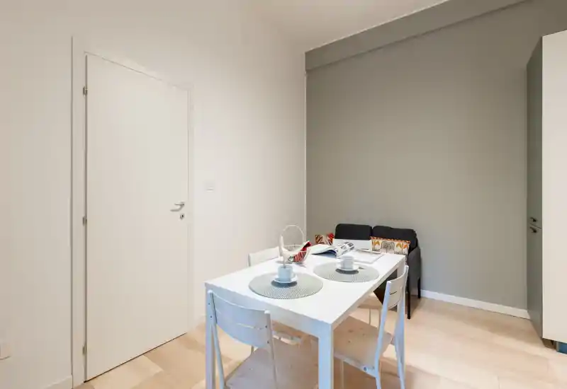 Zona pranzo luminosa e minimale con tavolo bianco apparecchiato per due e sedie semplici, adiacente a un piccolo angolo salotto; stile moderno e pulito ideale per appartamenti compatti.