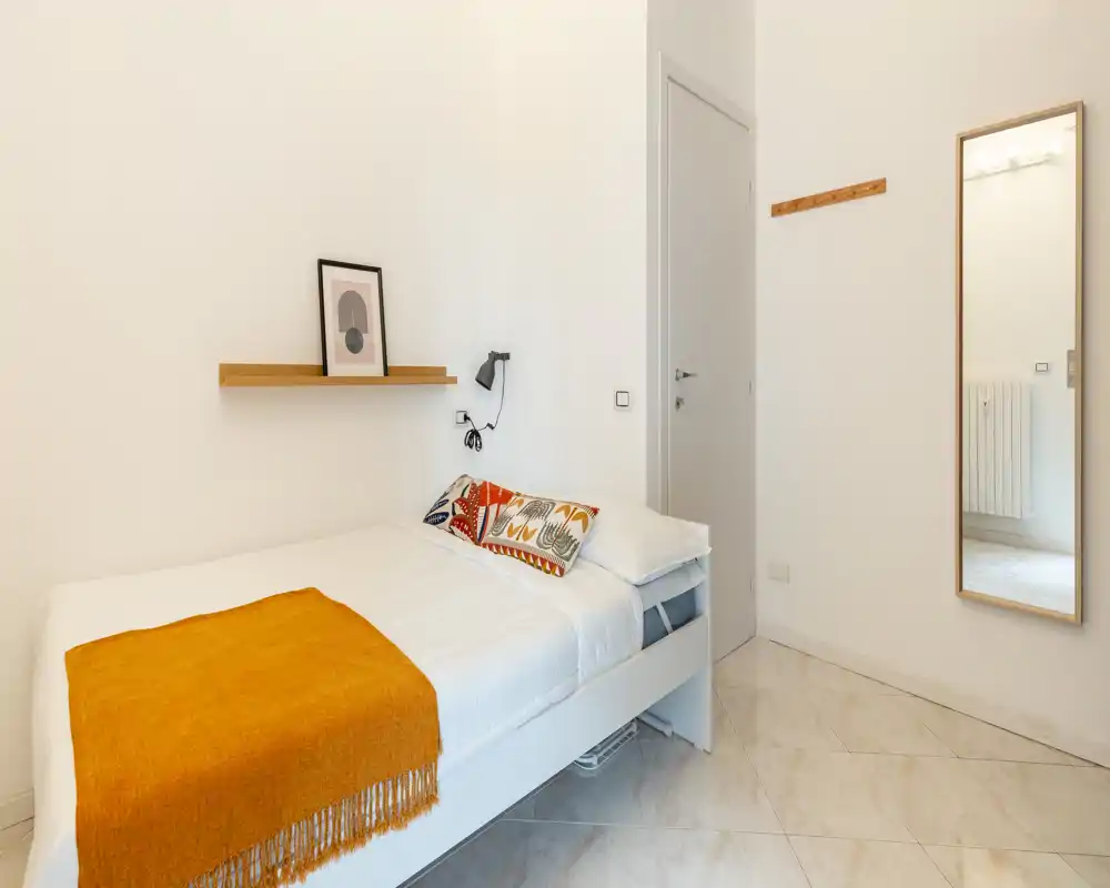 Compact, bright bedroom with a neatly made single bed, decorative cushions and a warm orange throw; simple shelf and wall lamp add practical style.