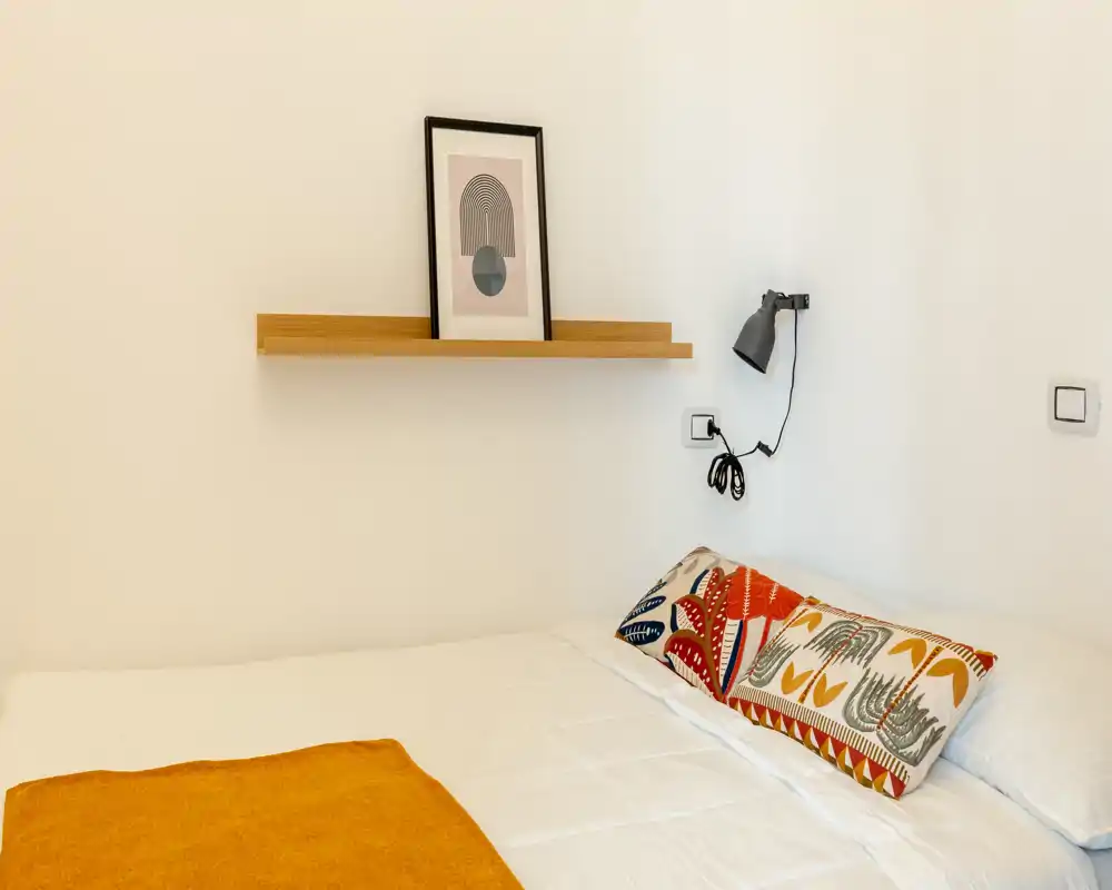 Sunny, minimalist bedroom corner with a neat double bed, patterned cushion and warm ochre throw against a clean white wall.