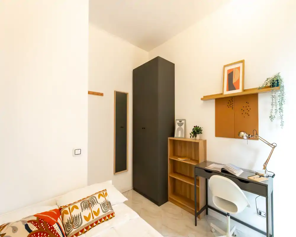 Bright, neatly staged bedroom corner with a partially visible bed, a tall dark wardrobe, open shelving and a small desk — a compact, functional room ideal for a single occupant or short-term rental.