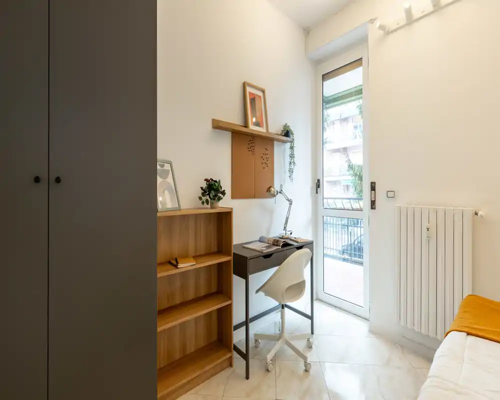 Small, well-lit bedroom corner with a single bed at the right edge, a compact study desk and chair by a balcony door, and built-in storage. The space feels tidy and functional for a student or single occupant.
