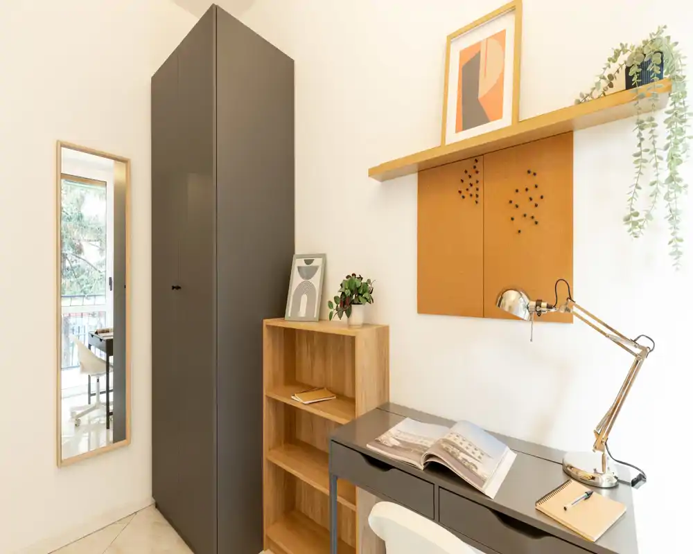 Bright, minimal bedroom corner with a tall wardrobe, shelving and a small desk ideal for remote work; neutral tones, framed art and plants create a calm, welcoming atmosphere.