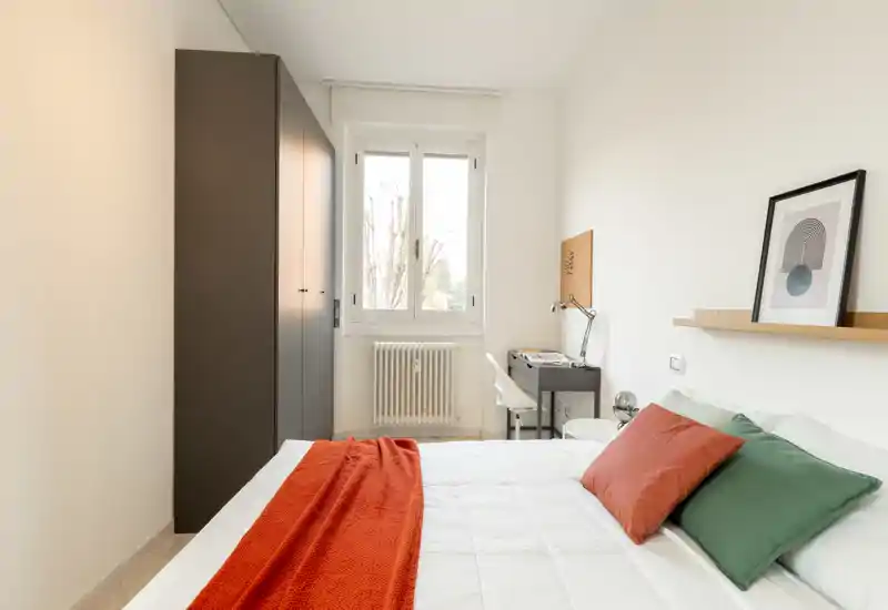 Camera da letto luminosa e minimal con letto matrimoniale con biancheria bianca, cuscini colorati e un plaid arancione. Armadio, scrivania e finestra con luce naturale completano lo spazio moderno e ordinato, perfetto per annunci in affitto.