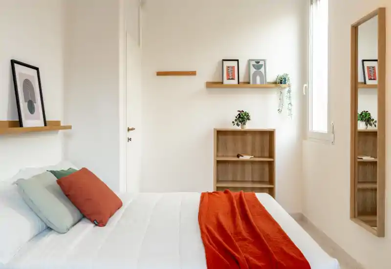 Camera da letto luminosa e minimal con letto ben rifatto, plaid arancione caldo e mensole in legno semplici che creano un'atmosfera calma e accogliente.
