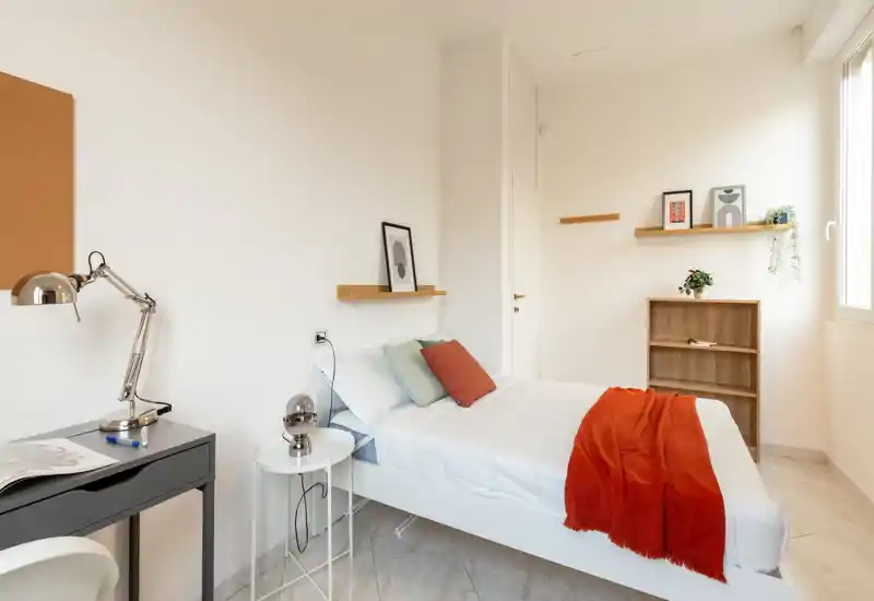 Camera da letto luminosa e minimal con letto singolo ben rifatto, cuscini colorati e un plaid rosso. La stanza ha una scrivania, un comodino e mensole con piante e quadretti, perfetta per un soggiorno accogliente.