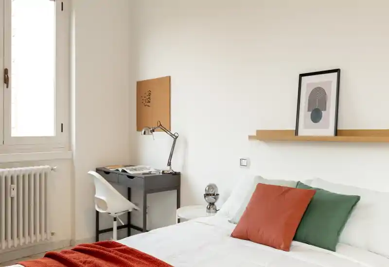 Camera da letto luminosa e minimal con letto matrimoniale rifinito in biancheria bianca, cuscini colorati e una coperta rossa. Un piccolo scrittoio con sedia vicino alla finestra offre una pratica postazione di lavoro alla luce naturale.