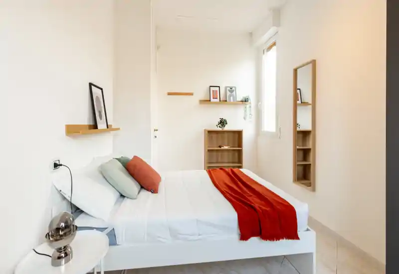 Camera da letto luminosa e minimal con un letto matrimoniale ben rifatto, cuscini colorati e un plaid rosso che aggiunge calore. Una finestra garantisce luce naturale, mentre mensole semplici e uno specchio completano l’aspetto pulito e arioso.