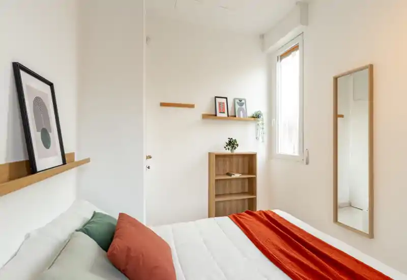 Camera da letto luminosa e minimale con letto ben rifatto, cuscini colorati e una coperta arancione. Mensole in legno, uno specchio alto e luce naturale dalla finestra creano un ambiente pulito e accogliente.