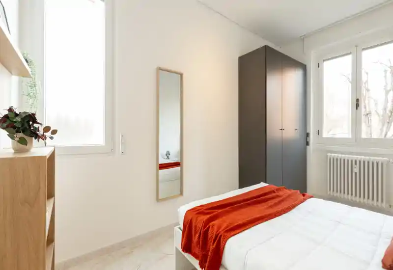 Camera da letto luminosa e minimale con letto ben rifatto in lenzuola bianche e un caldo plaid arancione; ampie finestre garantiscono molta luce naturale e un armadio alto offre spazio contenitivo.