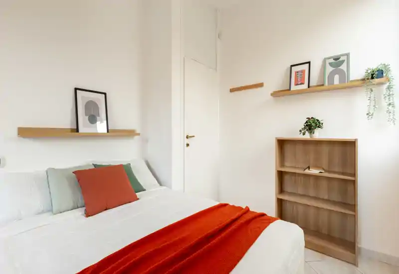 Camera da letto moderna e luminosa con letto matrimoniale rifinito, coperta arancione e mensole minimal; pareti neutre e luce naturale la rendono accogliente.