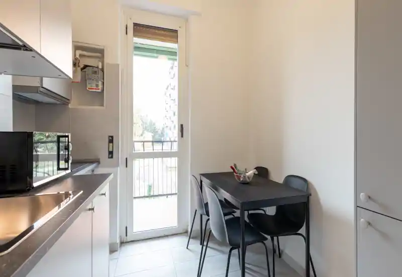 Cucina compatta e luminosa con un piccolo tavolo da pranzo per quattro persone. Pensili moderni, elettrodomestici sul piano e porta finestra sul balcone offrono luce naturale e una disposizione pratica.