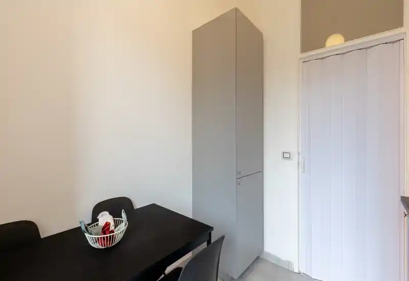 Angolo pranzo compatto con tavolo rettangolare scuro, quattro sedie e un cestino decorativo; pareti neutre e un armadietto alto creano un aspetto ordinato e minimalista.
