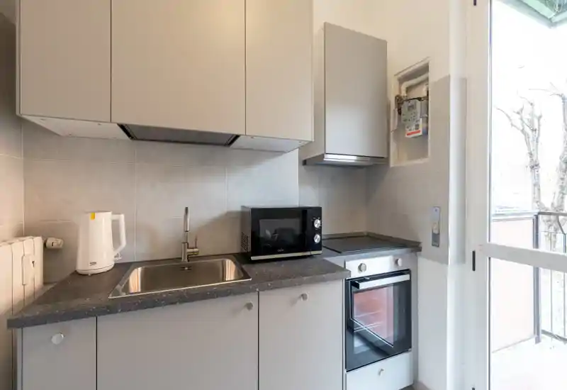 Cucina compatta e moderna con lavello, forno elettrico, piano cottura e microonde. Ambientazione luminosa con mobili neutri e porta sul balcone che assicura luce naturale.