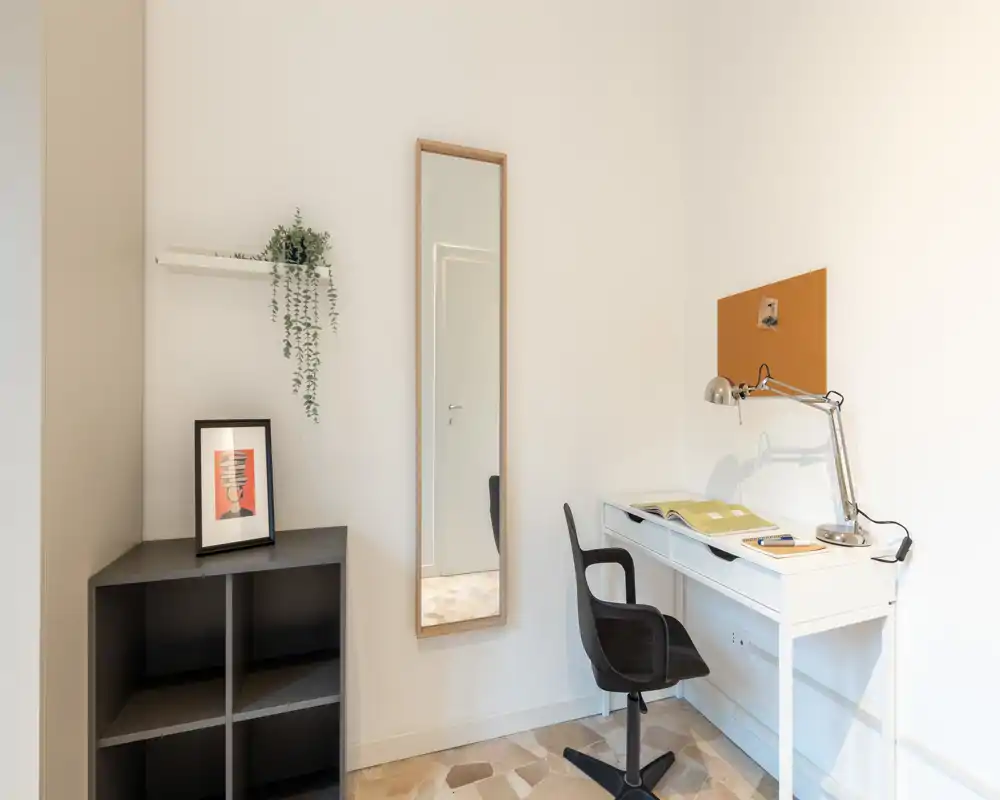 Coin bureau lumineux et minimaliste avec un bureau blanc, une lampe de bureau réglable et une chaise pivotante contre un mur épuré. Des étagères simples, un cadre et une grande glace apportent une touche décorative discrète.