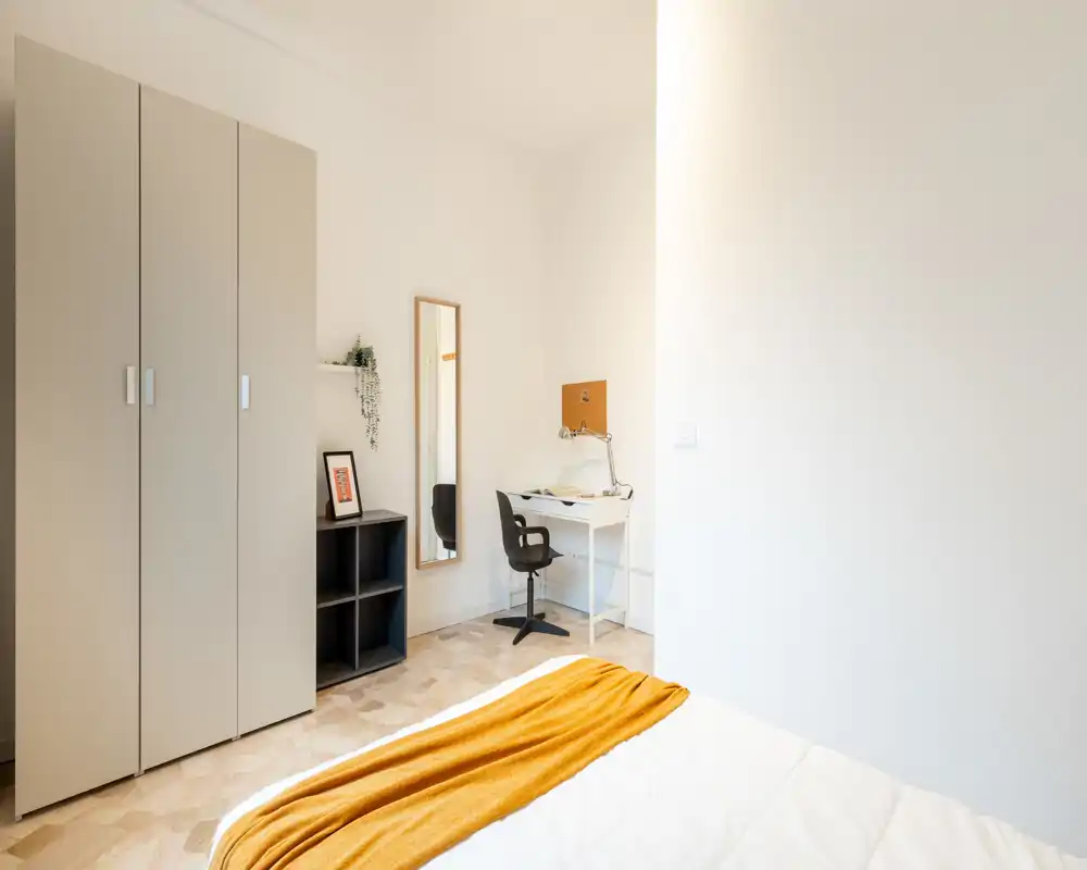 Chambre moderne et lumineuse avec lit bien fait et jeté ocre, armoire, miroir en pied et petit bureau parfait pour travailler ou étudier.