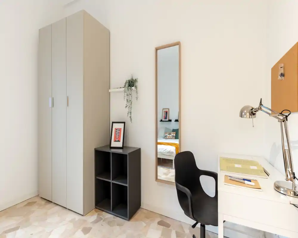 Coin de chambre lumineux et minimaliste avec armoire, bureau et chaise ; un miroir pleine‑longueur reflète le lit et donne de la profondeur. Style moderne et fonctionnel, adapté pour une annonce de chambre.