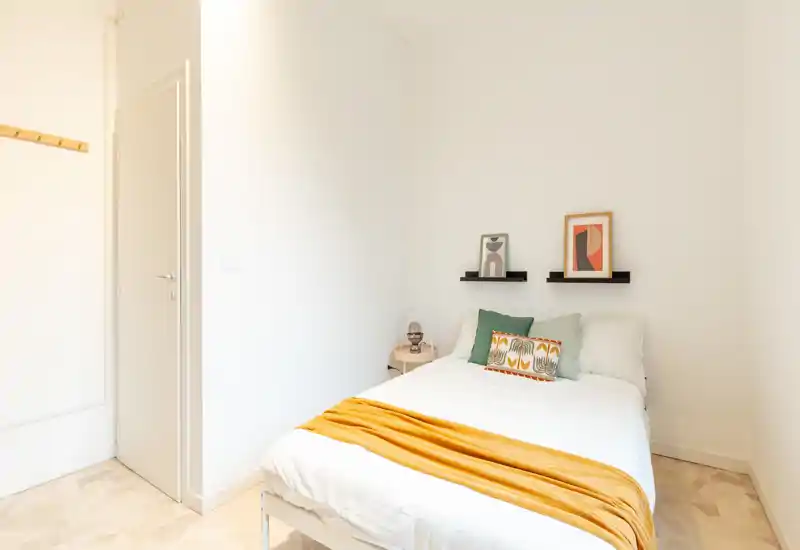 Chambre compacte et lumineuse avec un lit simple bien fait, plaid moutarde chaleureux et coussins décoratifs, petite table de chevet et décor minimaliste.