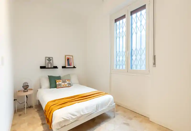 Chambre lumineuse et épurée avec un lit double bien fait, coussins décoratifs et un plaid moutarde. La lumière naturelle et le sol clair créent une ambiance aérée et apaisante.