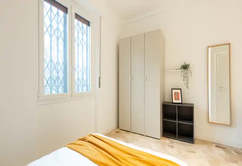 Chambre lumineuse et minimaliste avec une grande fenêtre, une armoire sobre et un plaid moutarde sur le lit qui apporte une touche chaleureuse.