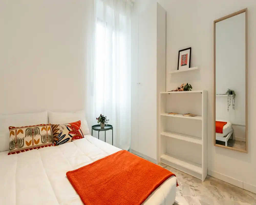Dormitorio moderno y luminoso con una amplia cama blanca, cojines decorativos y una manta naranja. La luz natural entra por las cortinas translúcidas; un espejo alto y estanterías aportan estilo y funcionalidad.